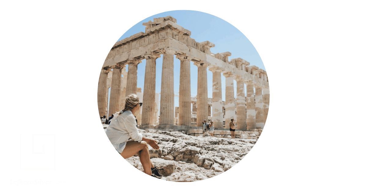 a woman seated outside an ancient Greek ruin and staring in wonder