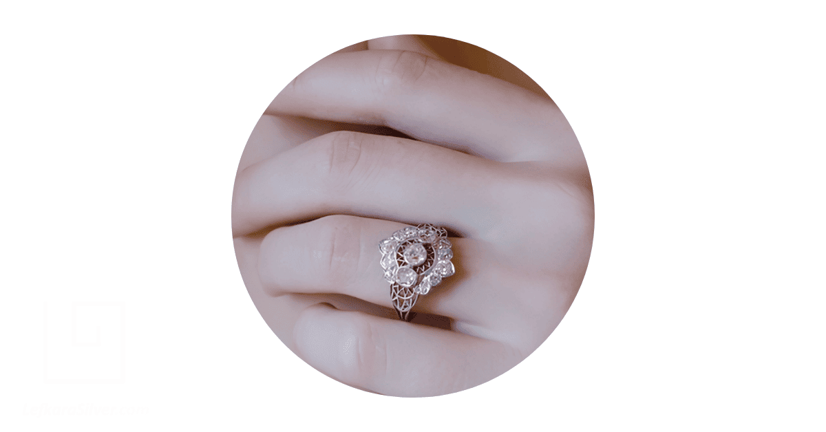 a woman's fingers with a silver filigree engagement ring sitting on the middle finger.