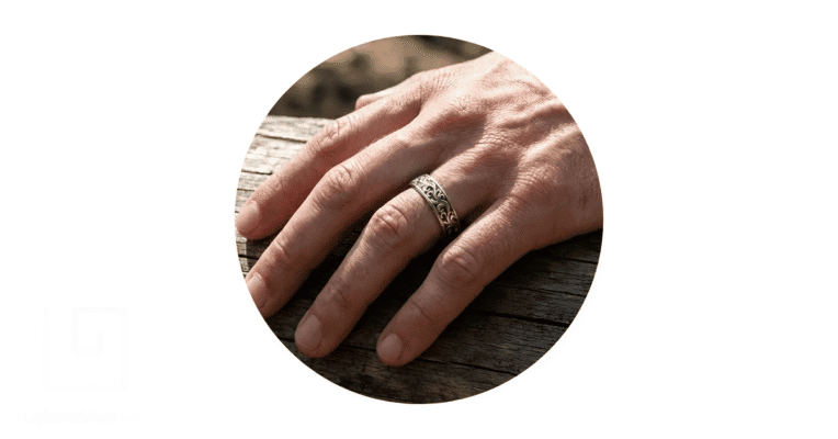 a man's hand on a wooden surface, with a filigree wedding band on his finger.